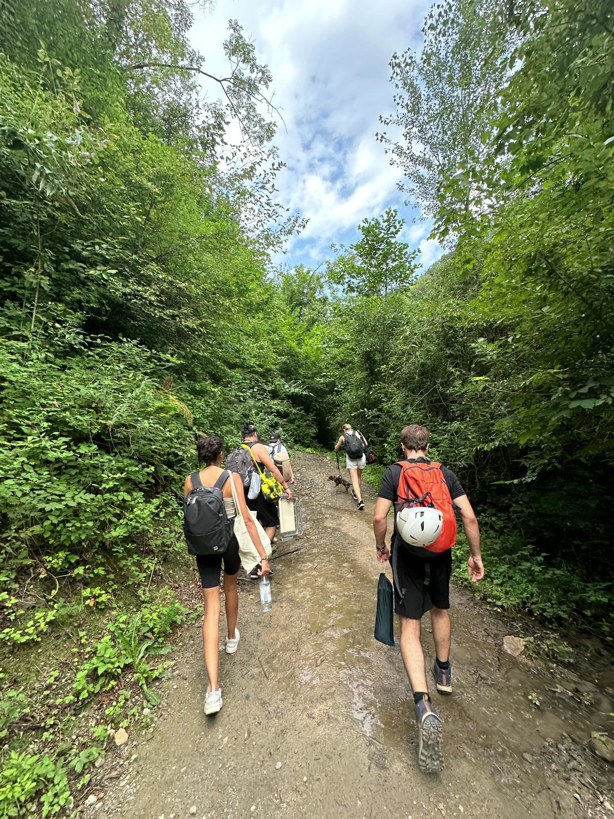 Friends hiking a muddy forest trail toward a crag, crash pad and helmet on their packs.