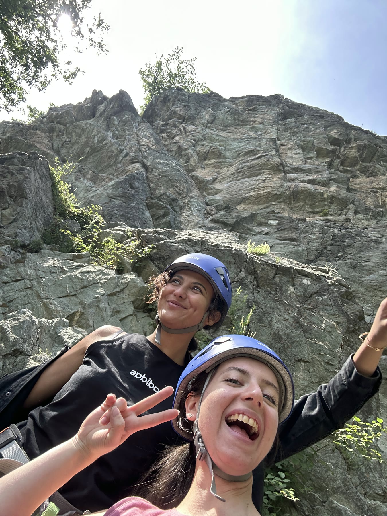 Two friends laughing in helmets at the base of a sunny crag.