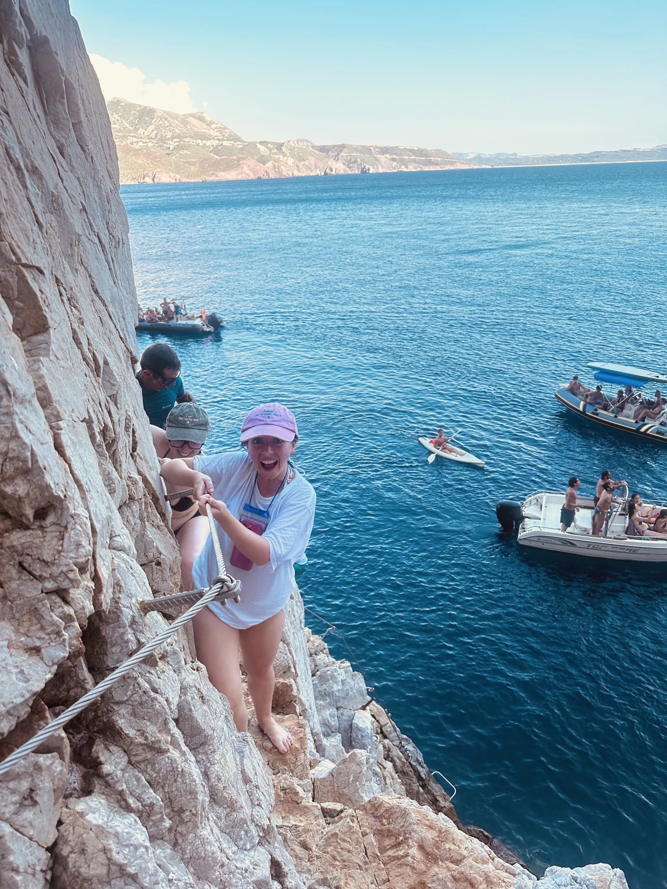 A climber gripping a via ferrata cable on a sea cliff, boats in the water below.